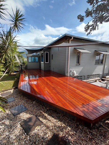 A freshly oiled merbau deck beside a weatherboard home with a shade sail and mountains behind